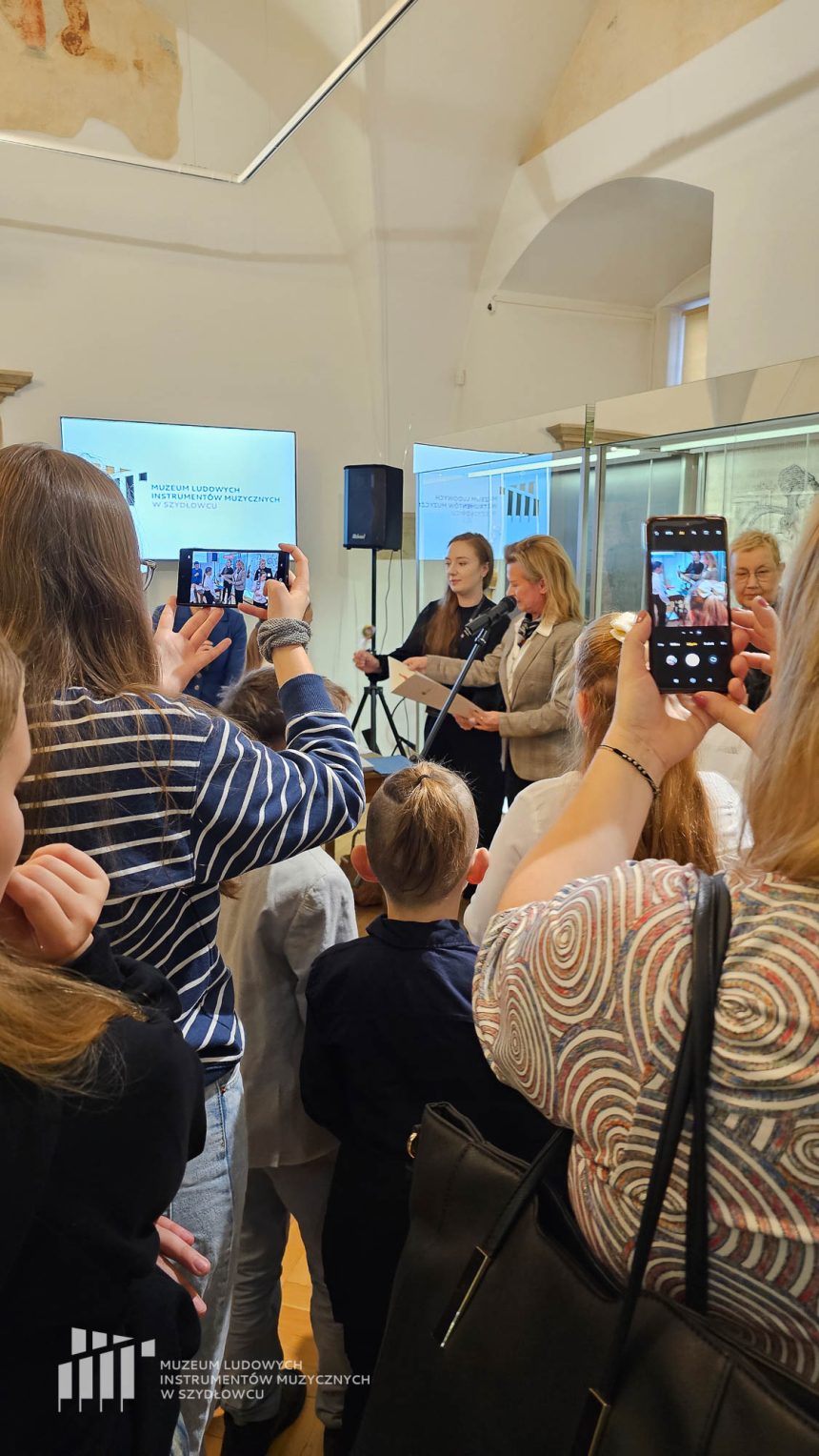 Dzieci stojące tyłem w sali muzealnej w komnatach zamku i patrzą na kobietę z blond włosami - Dyrektor Muzeum Instrumentów Anetę Oborny, która wręcza dyplomy dzieciom.