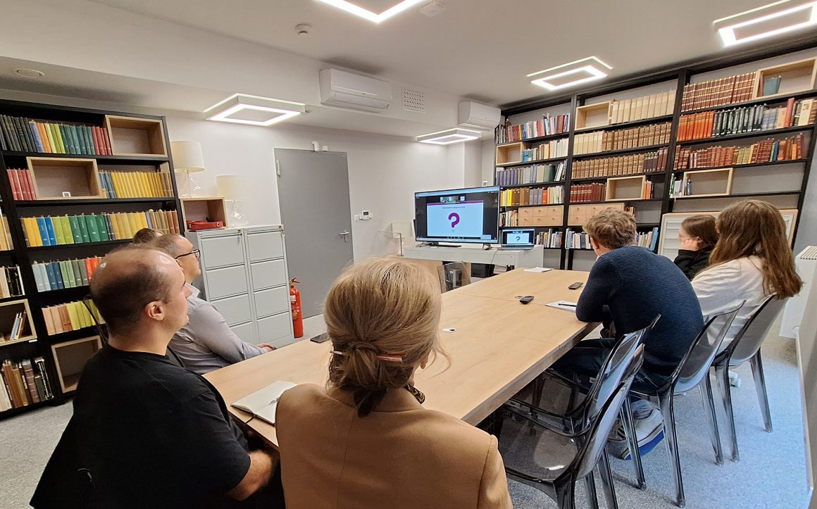 Ludzie siedzący przy długim stole w sali bibliotecznej oglądający prezentację na ekranie telewizyjnym.