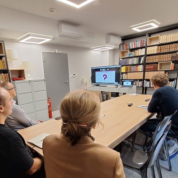 Ludzie siedzący przy długim stole w sali bibliotecznej oglądający prezentację na ekranie telewizyjnym.