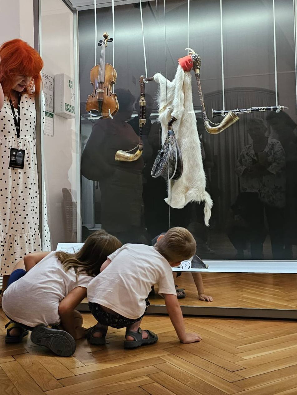 Przewodnik stojący przy ekspozycji oraz dwójka nachylających się dzieci podczas szukania lusterka w koźle weselnym białym.