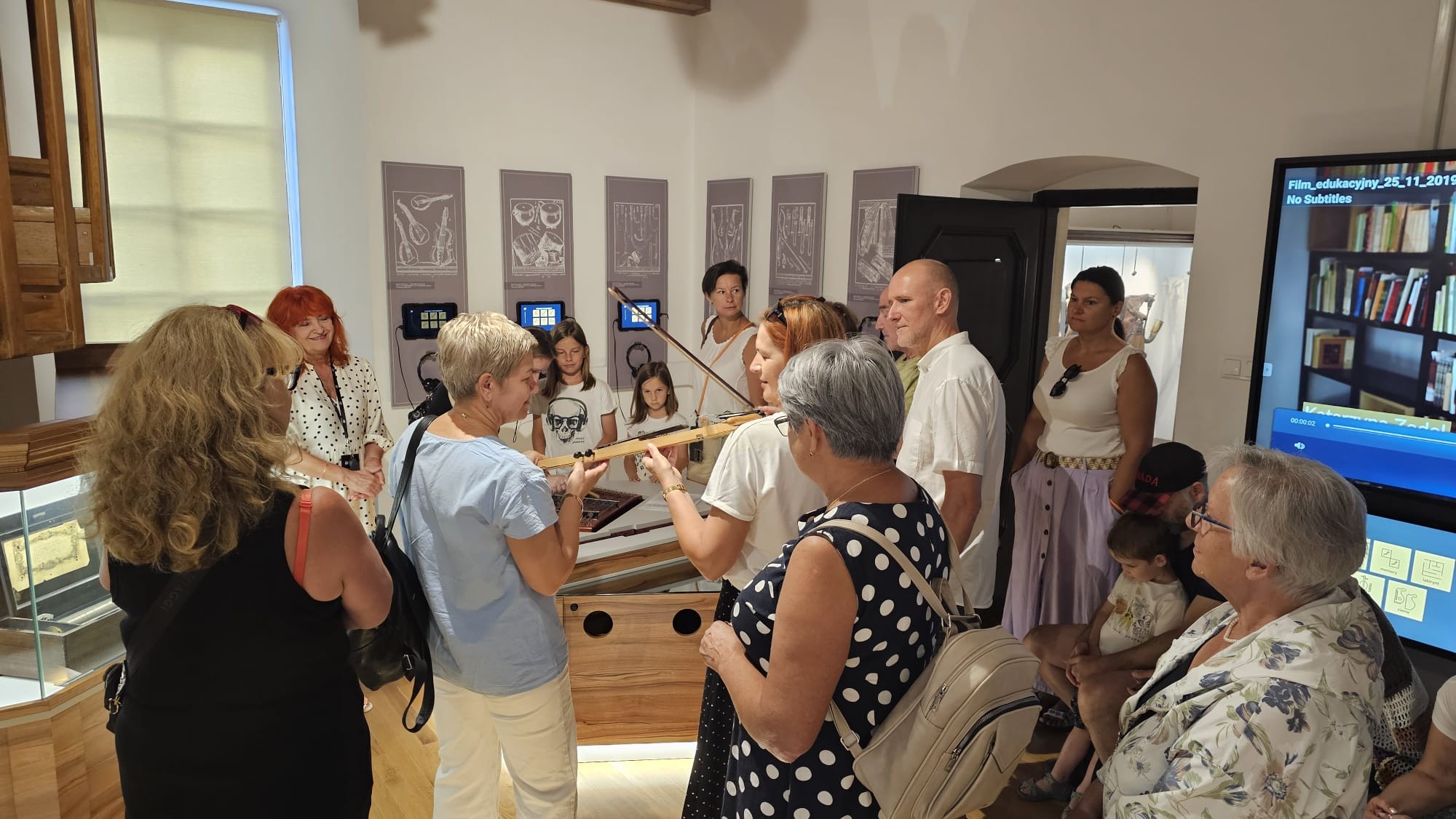 Grupa zwiedzających z przewodnikiem w Komnacie Intermezzo - Eksperymentarium Dźwięku przyglądająca się próbie grania na skrzypcach laskowych.