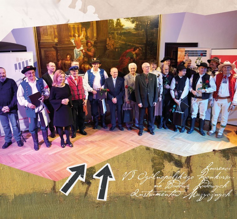 3-plansza_konkurs Zdjęcie. Grupa ludzi stojąca na scenie. Cześć z nich w strojach ludowych - laureaci VI Ogólnopolskiego Konkursu na Budowę Ludowych Instrumentów Muzycznych w 2022 roku