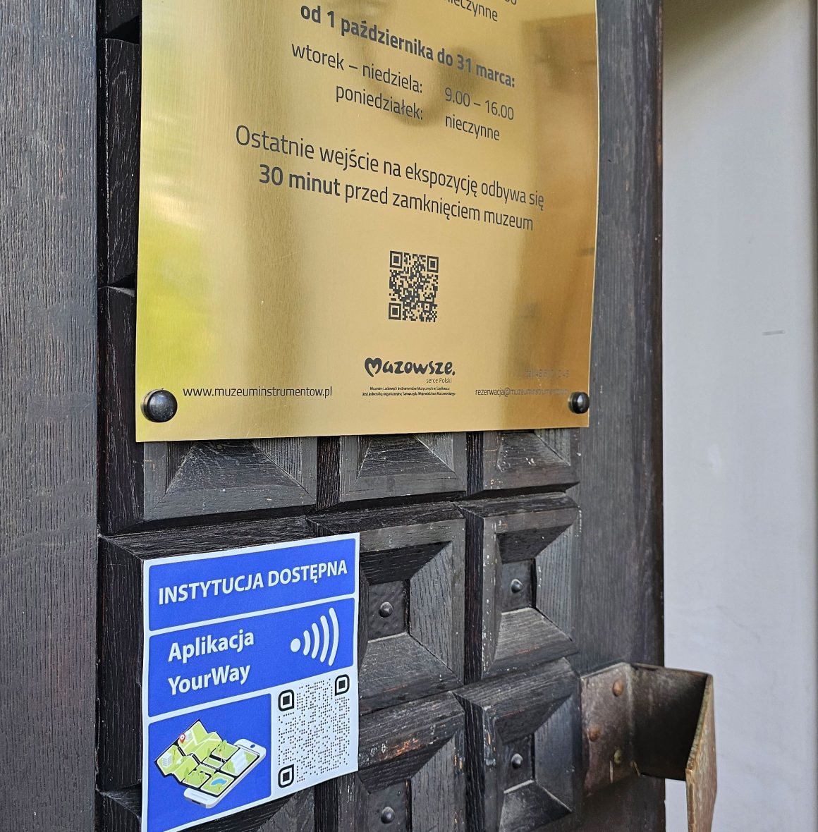 Zdjęcia. Drewniane ciemne drzwi. U góry złota tablica czarnymi napisami o godzinach otwarcia. Poniżej niebieska tabliczka z informacją o dostępności aplikacji YourWay.
