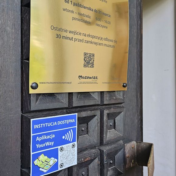 Zdjęcia. Drewniane ciemne drzwi. U góry złota tablica czarnymi napisami o godzinach otwarcia. Poniżej niebieska tabliczka z informacją o dostępności aplikacji YourWay.