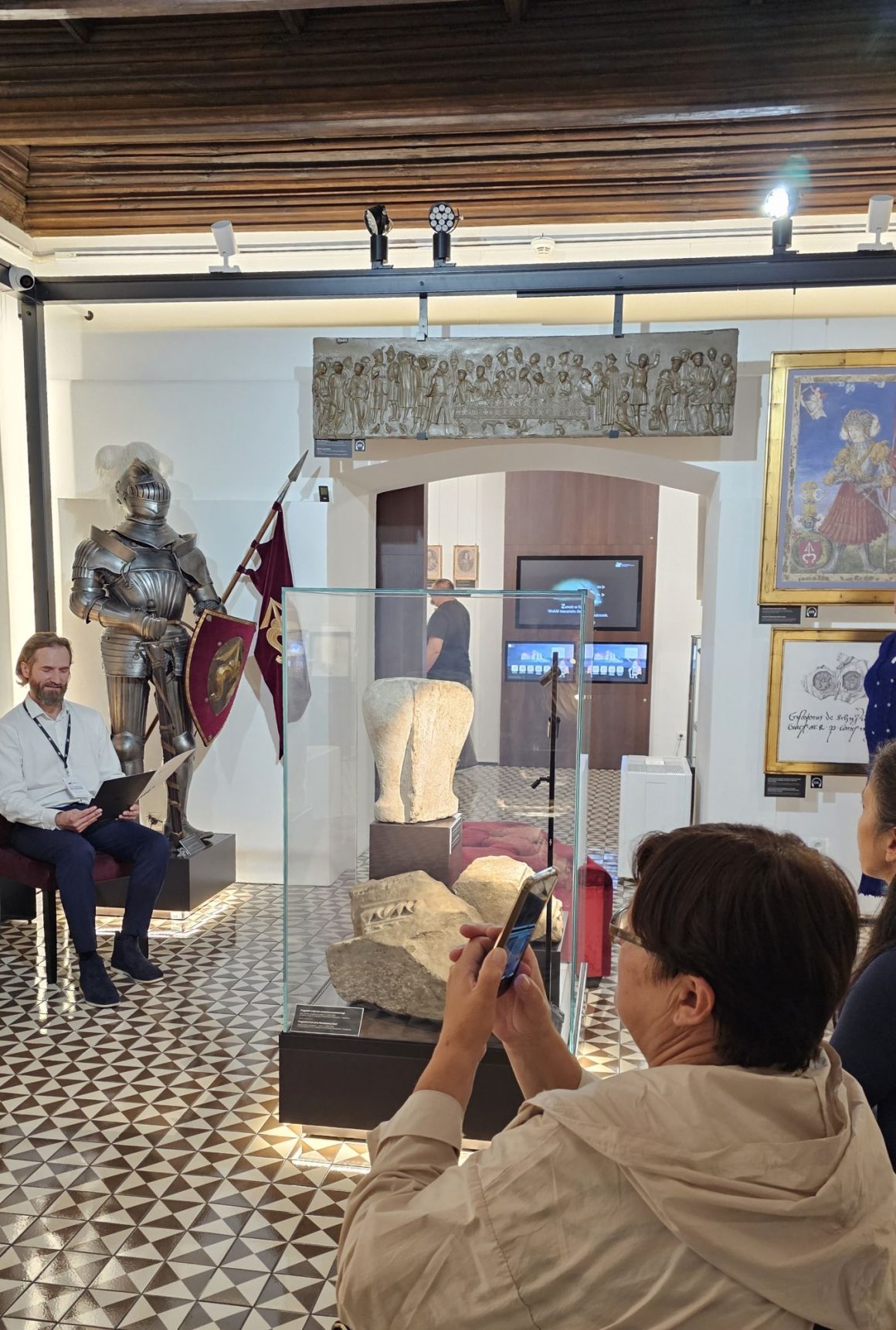 Narodowe Czytanie 2025 (2) Zdjęcie. W zamkowej komnacie po lewej stronie rycerska zbroja, na środku gablota z kamiennymi elementami. Przed zbroją na krześle siedzi mężczyzna i czyta. Po prawej stronie osoby patrzące na czytającego.
