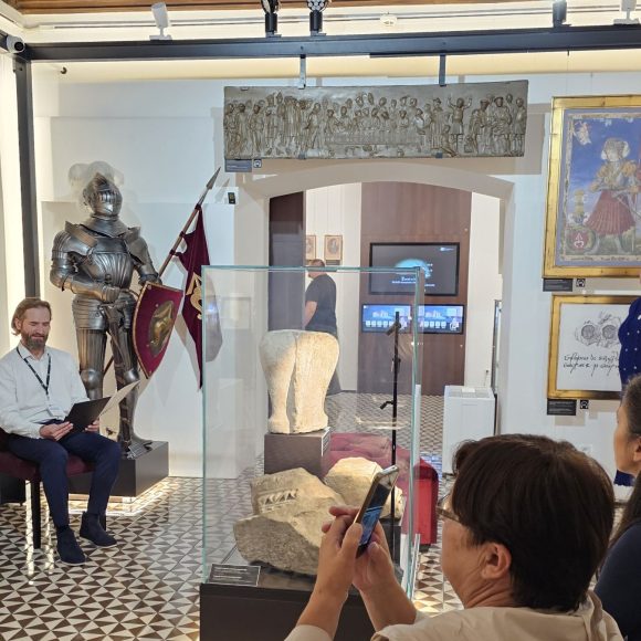 Narodowe Czytanie 2025 (2) Zdjęcie. W zamkowej komnacie po lewej stronie rycerska zbroja, na środku gablota z kamiennymi elementami. Przed zbroją na krześle siedzi mężczyzna i czyta. Po prawej stronie osoby patrzące na czytającego.