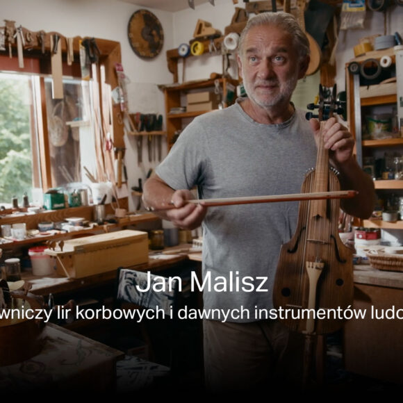 Zdjęcie twórcy lir korbowych i dawnych instrumentów ludowych - Jana Malisza w swojej pracowni podczas gry na skrzypcach. W tle instrumenty i narzędzia rzemieślnicze. Ubrany jest w szarą koszulkę.
