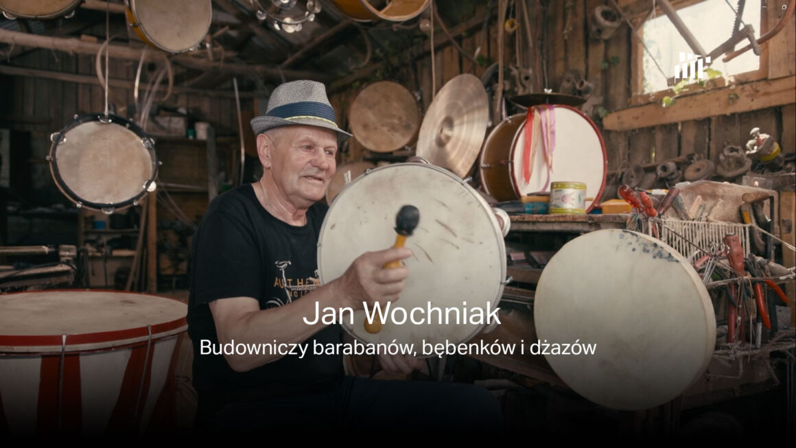 Zdjęcie twórcy barabanów, bębenków i dżazów - Jana Wochniaka w swojej pracowni podczas gry na bębenku. W tle instrumenty i narzędzia rzemieślnicze. Na głowie ma kapelusz.