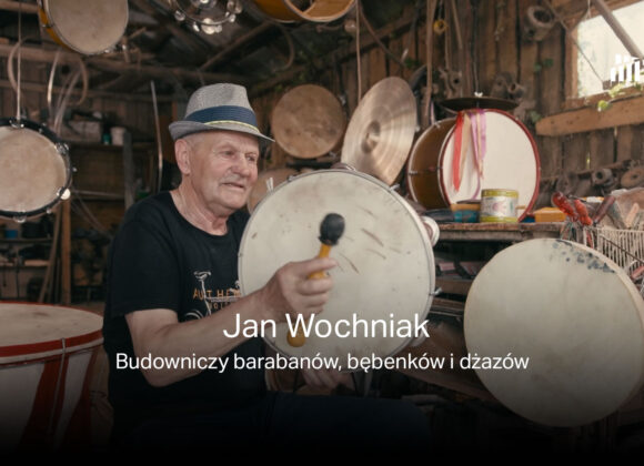 Zdjęcie twórcy barabanów, bębenków i dżazów - Jana Wochniaka w swojej pracowni podczas gry na bębenku. W tle instrumenty i narzędzia rzemieślnicze. Na głowie ma kapelusz.