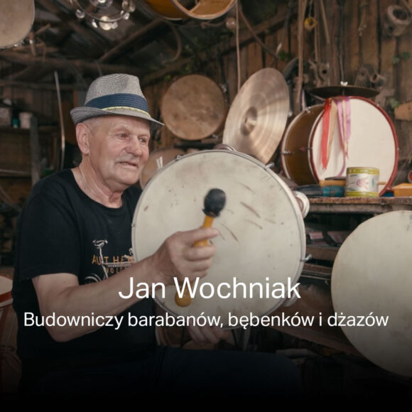 Zdjęcie twórcy barabanów, bębenków i dżazów - Jana Wochniaka w swojej pracowni podczas gry na bębenku. W tle instrumenty i narzędzia rzemieślnicze. Na głowie ma kapelusz.