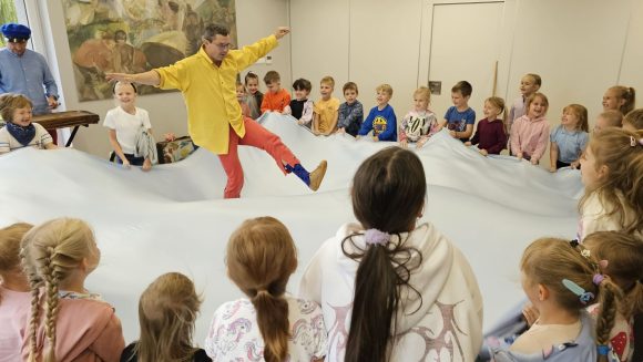 Aktorzy z teatrzyku „Słuchaj uchem” oraz dziecięca publiczność podczas spektaklu „Kusy Janek”. Dzieci trzymają i ruszają brzegi błękitnego materiału imitując morskie fale, a aktor stoi na środku i odgrywa swoją rolę.