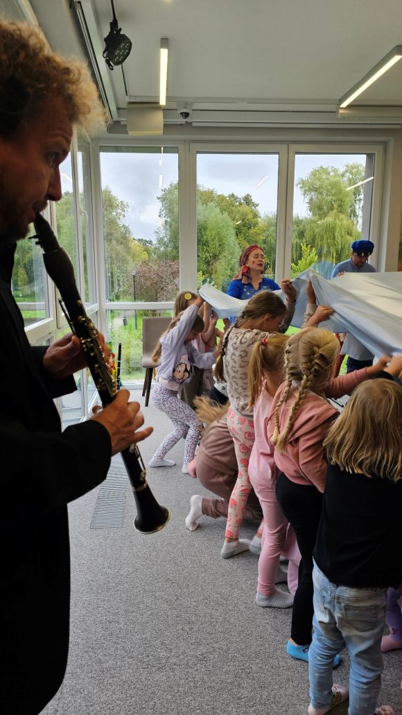 Aktorzy z teatrzyku „Słuchaj uchem” oraz dziecięca publiczność podczas spektaklu „Kusy Janek”. Część dzieci uczestniczy w zabawie tanecznej, reszta siedzi i ogląda spektakl.
