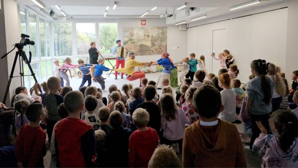 Aktorzy z teatrzyku „Słuchaj uchem” oraz dziecięca publiczność podczas spektaklu „Kusy Janek”. Część dzieci uczestniczy w zabawie tanecznej, reszta siedzi i ogląda spektakl.