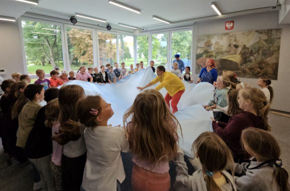 Aktorzy z teatrzyku „Słuchaj uchem” oraz dziecięca publiczność podczas spektaklu „Kusy Janek”. Dzieci trzymają i ruszają brzegi błękitnego materiału imitując morskie fale, a aktor stoi na środku i odgrywa swoją rolę.