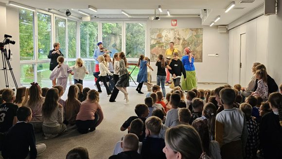 Aktorzy z teatrzyku „Słuchaj uchem” oraz dziecięca publiczność podczas spektaklu „Kusy Janek”. Część dzieci uczestniczy w zabawie tanecznej, reszta siedzi i ogląda spektakl.