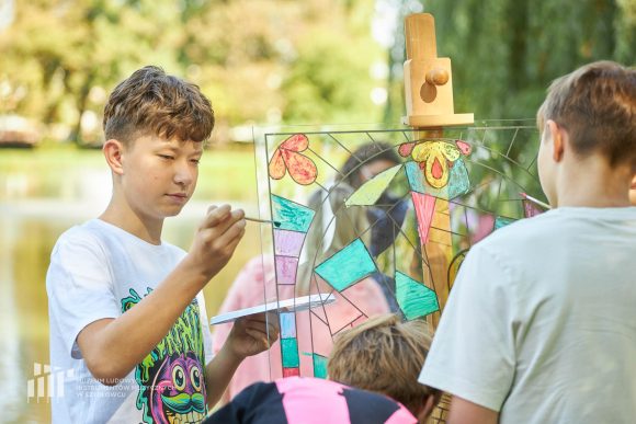 Na pierwszym planie uczniowie Publicznej Szkoły Podstawowej nr 2 im. Jana Pawła II w Szydłowcu podczas zajęć artystycznych „Witraż w plenerze”. W tle fosa przy zamku.