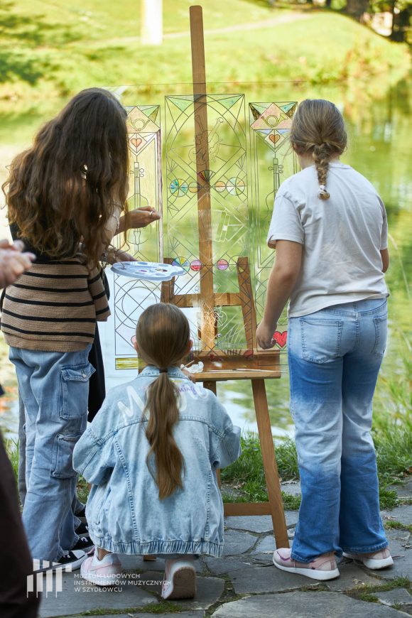 Na pierwszym planie uczniowie Publicznej Szkoły Podstawowej nr 2 im. Jana Pawła II w Szydłowcu podczas zajęć artystycznych „Witraż w plenerze”. W tle fosa przy zamku.