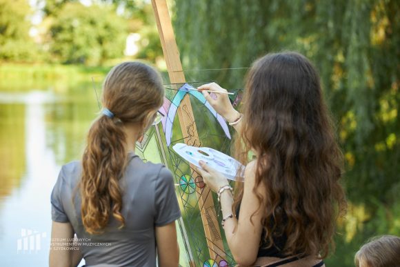 Na pierwszym planie uczniowie Publicznej Szkoły Podstawowej nr 2 im. Jana Pawła II w Szydłowcu podczas zajęć artystycznych „Witraż w plenerze”. W tle fosa przy zamku.