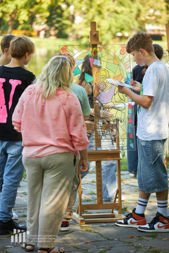 Na pierwszym planie uczniowie Publicznej Szkoły Podstawowej nr 2 im. Jana Pawła II w Szydłowcu podczas zajęć artystycznych „Witraż w plenerze”. W tle fosa i fragment zamku.
