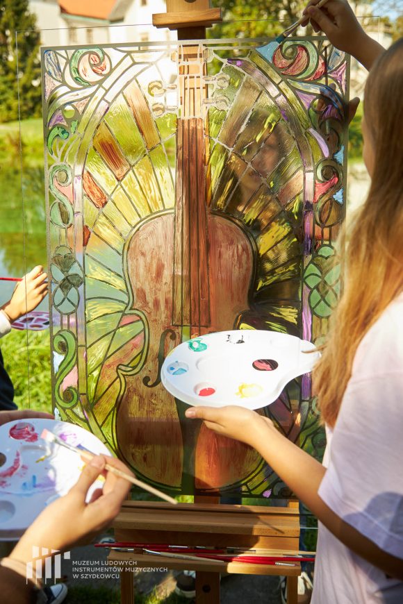 Na pierwszym planie uczniowie Publicznej Szkoły Podstawowej nr 2 im. Jana Pawła II w Szydłowcu podczas zajęć artystycznych „Witraż w plenerze”. W tle fosa i fragment zamku.