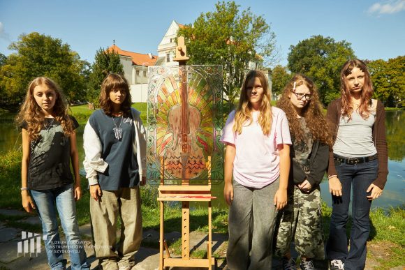 Na pierwszym planie uczniowie Publicznej Szkoły Podstawowej nr 2 im. Jana Pawła II w Szydłowcu prezentujący swoją pracę podczas zajęć artystycznych „Witraż w plenerze”. W tle fosa i fragment zamku.