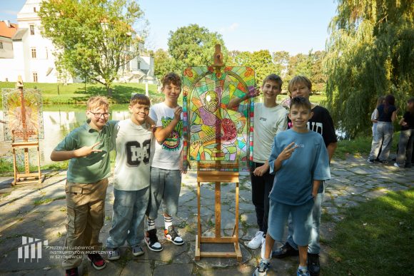 Na pierwszym planie uczniowie Publicznej Szkoły Podstawowej nr 2 im. Jana Pawła II w Szydłowcu prezentujący swoją pracę podczas zajęć artystycznych „Witraż w plenerze”. W tle fosa i fragment zamku.