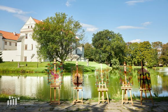 Na pierwszym planie imitacja pięciu witraży, efekt pracy uczniów Publicznej Szkoły Podstawowej nr 2 im. Jana Pawła II w Szydłowcu namalowanych podczas zajęć artystycznych „Witraż w plenerze”. W tle fosa i fragment zamku.