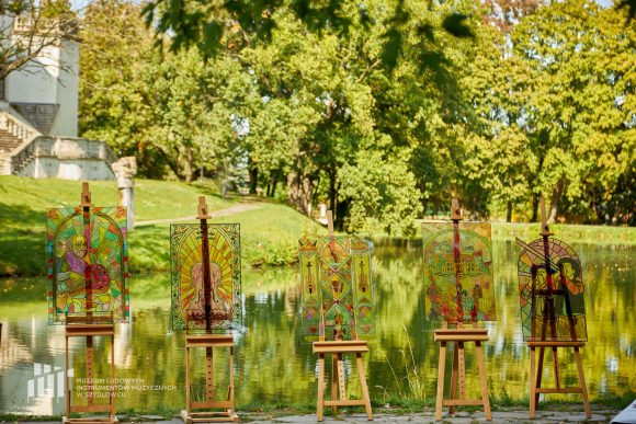 Na pierwszym planie imitacja pięciu witraży, efekt pracy uczniów Publicznej Szkoły Podstawowej nr 2 im. Jana Pawła II w Szydłowcu namalowanych podczas zajęć artystycznych „Witraż w plenerze”. W tle fosa i fragment zamku.