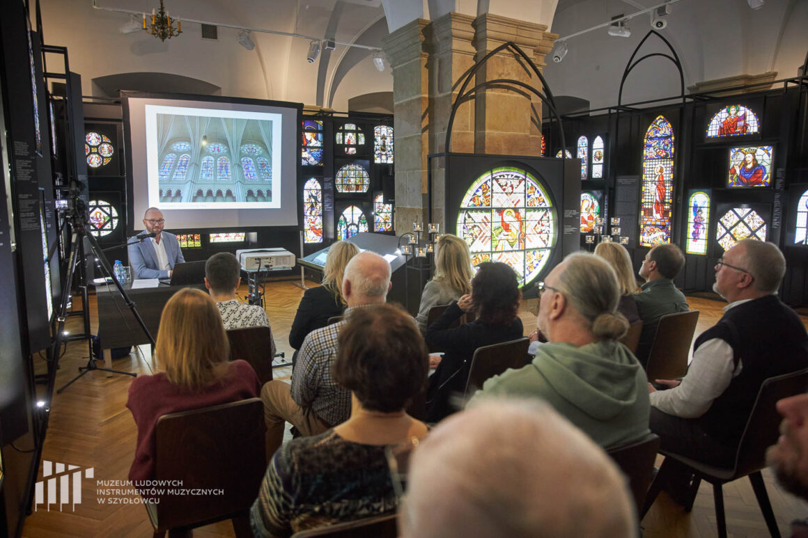Na zdjęciu 12 osób widzianych od tyłu podczas spotkania „Niebiańskie Jeruzalem. Witraż. Historia, technika, przykłady”. Siedzą na krzesełkach w otoczeniu wystawy czasowej „Muzyka światła. Witraże. Od artefaktu do metafizyki”. Na przeciwko nich siedzi prowadzący dr Adam Włodarczyk mówiący do mikrofonu. Po prawej lightbox z podświetlaną reprodukcją witrażu św. Cecylii z kaplicy pw. św. Józefa w Sanktuarium Miłosierdzia Bożego w Krakowie-Łagiewnikach oraz inne lightboxes wystawy.