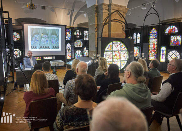 Na zdjęciu 12 osób widzianych od tyłu podczas spotkania „Niebiańskie Jeruzalem. Witraż. Historia, technika, przykłady”. Siedzą na krzesełkach w otoczeniu wystawy czasowej „Muzyka światła. Witraże. Od artefaktu do metafizyki”. Na przeciwko nich siedzi prowadzący dr Adam Włodarczyk mówiący do mikrofonu. Po prawej lightbox z podświetlaną reprodukcją witrażu św. Cecylii z kaplicy pw. św. Józefa w Sanktuarium Miłosierdzia Bożego w Krakowie-Łagiewnikach oraz inne lightboxes wystawy.