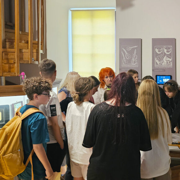 Uczniowie podczas zwiedzania w sali Eksperymentarium Dźwięku, skupieni wokół stolika z cymbałami i skrzypcami laskowymi słuchający przewodnika muzealnego.