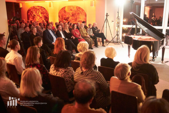 Na zdjęciu po lewej fortepian oraz ukryty za nim pianista podczas recitalu fortepianowego w sali koncertowej budynku administracyjno-biurowego Muzeum Ludowych Instrumentów Muzycznych w Szydłowcu. Po lewej w głównej części sali konferencyjnej oraz na pierwszym planie zdjęcia w mniejszym segmencie sali zgromadzona publiczność zasłuchana w muzyce.