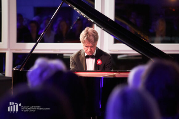 Na zdjęciu zbliżenie na grającego pianistę Pawła Kowalskiego i fortepian podczas recitalu fortepianowego w sali koncertowej budynku administracyjno-biurowego Muzeum Ludowych Instrumentów Muzycznych w Szydłowcu. W tle zdjęcia okna, przez które widać wieczór, a na pierwszym planie cienie głów publiczności w niebieskiej poświacie.