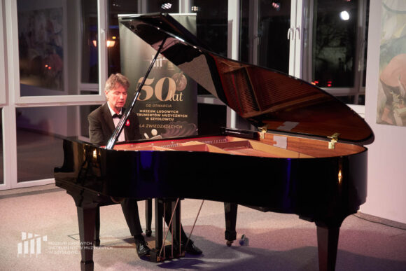 Na zdjęciu zbliżenie na grającego pianistę Pawła Kowalskiego i fortepian podczas recitalu fortepianowego w sali koncertowej budynku administracyjno-biurowego Muzeum Ludowych Instrumentów Muzycznych w Szydłowcu. W tle zdjęcia okna, przez które widać wieczór, a przed oknami rollup z informacją o 50 latach otwarcia muzeum dla zwiedzających.