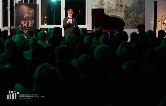 W tle oświetlony przez lampy pianista Paweł Kowalski przemawiający do publiczności, zapowiadający swój występ podczas recitalu fortepianowego w Muzeum Ludowych Instrumentów Muzycznych w Szydłowcu. Na pierwszym planie nieoświetlona publiczność słuchająca jego wystąpienia.