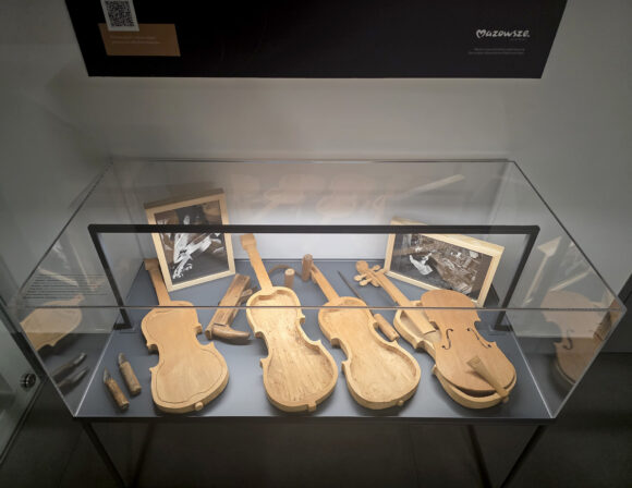 Zdjęcie przedstawia drewniane pudło rezonansowe skrzypiec w trakcie budowy, w czerech etapach – widoczna jest górna płyta z wyciętymi otworami rezonansowymi, obok znajdują się narzędzia stolarskie używane do formowania i wykończenia instrumentu.