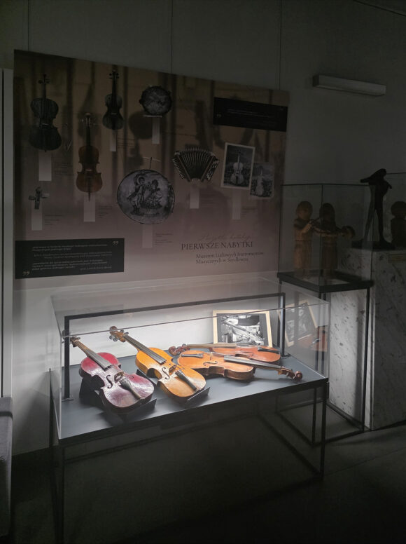 Pokazana w półmroku i odświetlona gablota wystawy „Początki kolekcji. Pierwsze nabytki Muzeum Ludowych Instrumentów Muzycznych w Szydłowcu” – wewnątrz skrzypce wykonane i klejone przez różnych twórców, a nad gablotą tablica informacyjna z reprodukcjami pierwszych fotografii dokumentujących początki zbiorów i pierwsze wystawy.
