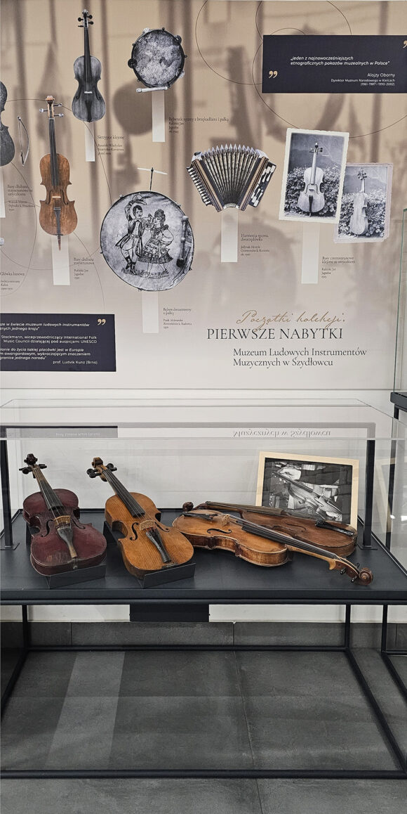 Gablota wystawy „Początki kolekcji. Pierwsze nabytki Muzeum Ludowych Instrumentów Muzycznych w Szydłowcu” – wewnątrz skrzypce wykonane i klejone przez różnych twórców, a nad gablotą tablica informacyjna z reprodukcjami pierwszych fotografii dokumentujących początki zbiorów i pierwsze wystawy.