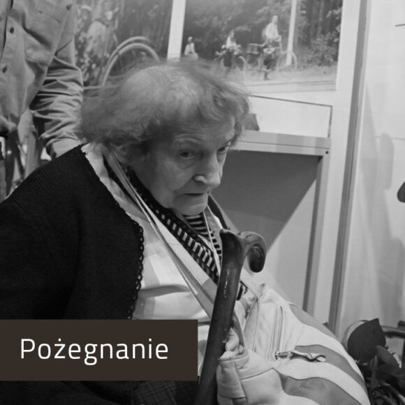 Postać starszej kobiety na czarnobiałym zdjęciu. Siedzi we wnętrzu muzeum z drewnianą laską i jasną torbą przewieszoną przez ramię. Ma na sobie ciemny sweter i białą koszulę z apaszką w kontrastowe pasy.