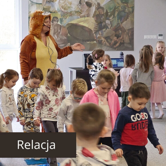 Animatorka w żółto brązowym stroju lwa z mikrofonem przy twarzy. Obok gromada dzieci biegających gęsiego po matach sensorycznych.