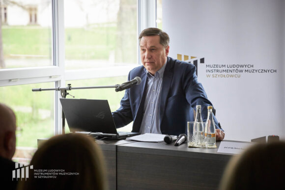 Prelegent siedzi przy stoliku przy mikrofonie przed publicznością. Na stoliku stoi woda w butelkach i szklanki, laptop, mokrofony i materiały. Za nim rollup Muzeum w Szydłowcu i okna ze światłem dziennym.