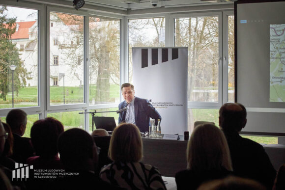 Prelegent siedzi przy stoliku przy mikrofonie przed publicznością. Na stoliku stoi woda w butelkach i szklanki. Za nim rollup Muzeum w Szydłowcu i okna ze światłem dziennym z widokiem na zamek.