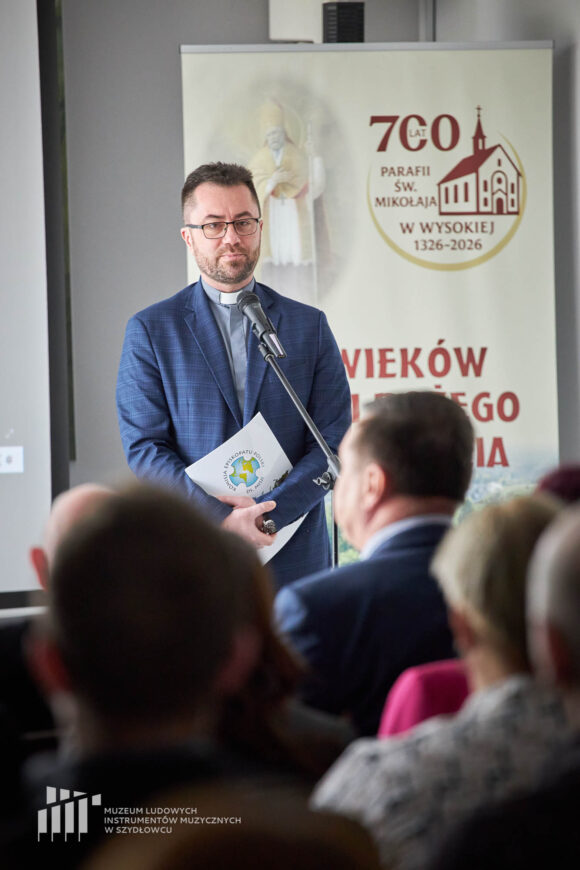 Na tle rollupu "700 lat Parafii Św. Mikołaja w Wysokiej" stoi ksiądz przed mikrofonem i przemawia do siedzącej publiczności. W dłoniach trzyma papierową teczkę.