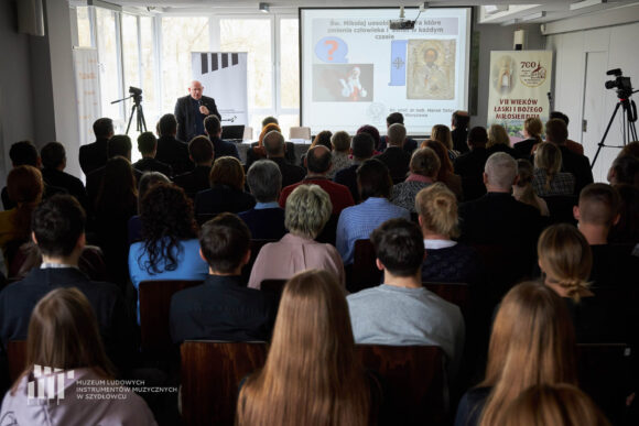W oddali stoi ksiądz i przemawia przez mikrofon, przed nim publiczność, za nimi na ekranie prezentacja multimedialna oraz trzy rollupy po bokach.
