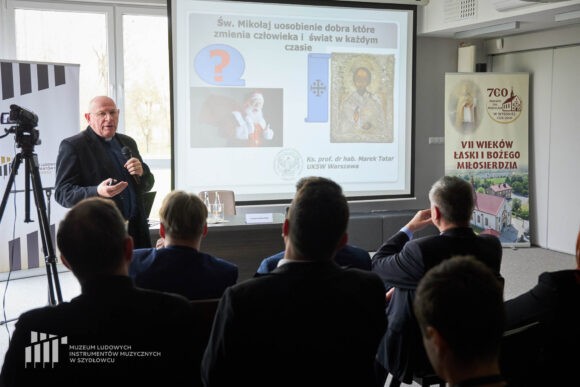 Po lewej ksiądz w granatowej koszuli z koloratką w czarnej marynarce stoi i przemawia przez mikrofon, który trzyma w ręku. Przed nim siedzi publiczność, obok stoi statyw.