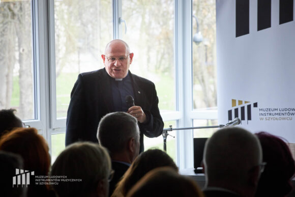 Ksiądz w granatowej koszuli z koloratką w czarnej marynarce stoi i przemawia przez mikrofon, który trzyma w ręku. Przed nim siedzi publiczność.