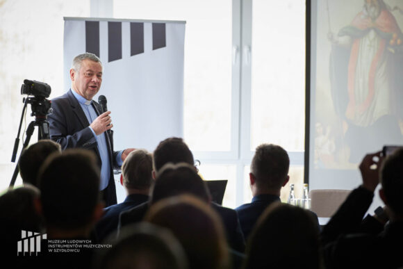 Po lewej ksiądz w granatowej koszuli z koloratką w czarnej marynarce stoi i przemawia przez mikrofon, który trzyma w ręku. Przed nim siedzi publiczność.