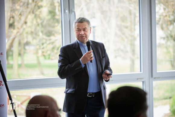 Ksiądz w granatowej koszuli z koloratką w czarnej marynarce stoi i przemawia przez mikrofon, który trzyma w ręku.