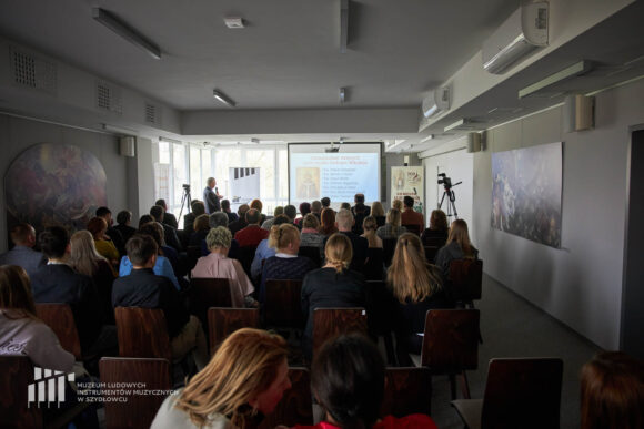 Zdjęcie z końca sali na publiczność, prowadzącego i prezentację.