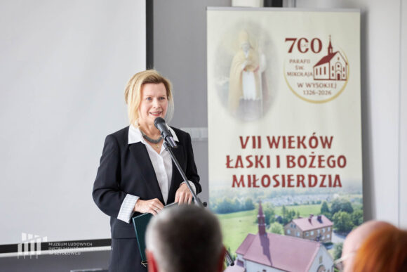 Uśmiechnięta dyrektor muzeum rozpoczynająca sympozjum historyczne. Obok widoczny jest rollup "700 lat parafii św. Mikołaja w Wysokiej".